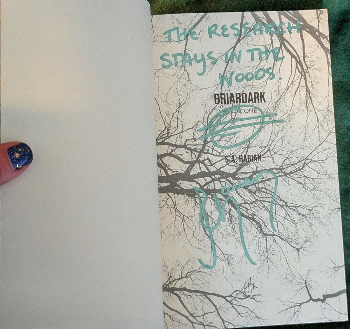 Briardark's signed title page featuring tree branches and text, held by a hand with blue nail polish.